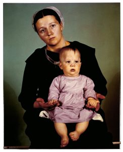 Dark-haired woman in a dark dress holds in her lap a child in a lavender dress.