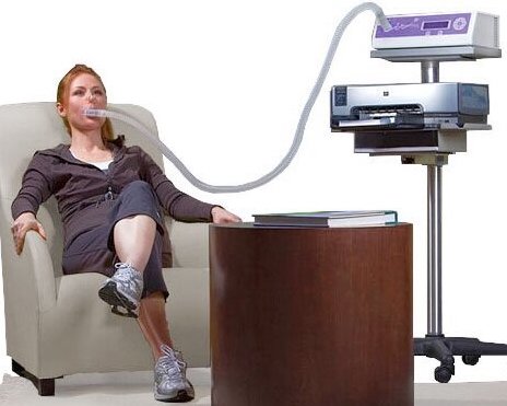 Woman sits in an armchair with legs crossed and a tube in her mouth that leads to a medical machine to measure oxygen.