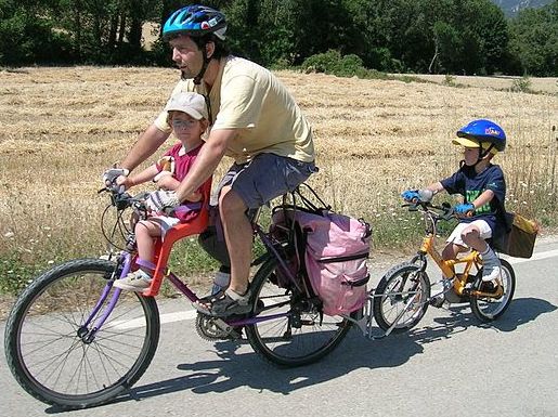 A father bikes with a young child in front of him and another young child on their own bike behind him.