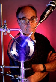 Dark-haired balding man with thick eyebrows and large glasses crosses his arms while staring forward past a scientific contraption involving a ring stand, a dark sphere with a purple tube going through its middle and something being injected from the upper right.