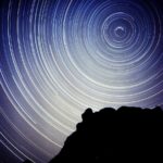 Time lapse photo showing multiple circles of bright light in the night sky. A rocky outcropping completely in shadow is in the foreground.